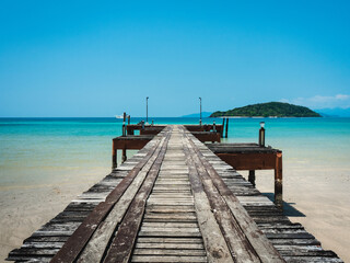 Scenic view of long rustic wooden pier over crystal clear turquoise water and white sand beach of Koh Mak Island with Koh Kham Island at horizon, Trat, Thailand.