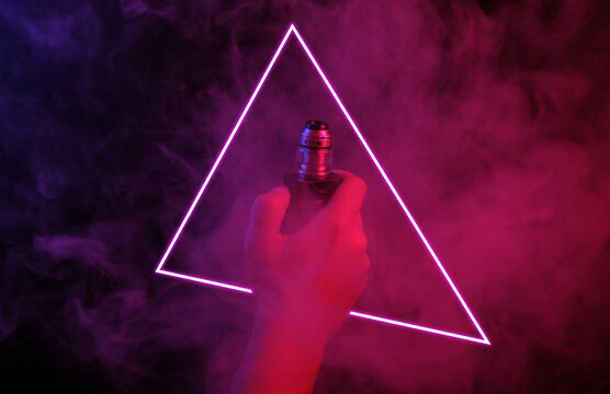Hand holds electronic cigarette or vaping device in glowing triangle and dense smoke with pink blue neon light.