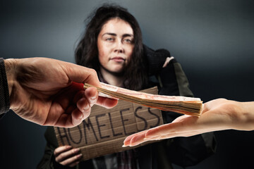 Man gives a stack of cash to woman, hands close up. Defocused disheveled woman holds a cardboard sign with the text homeless on the background. The concept of helping the homeless and vagrants