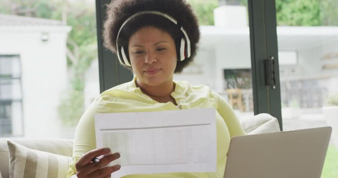 Video of happy plus size african american woman with headphones sitting on sofa with laptop