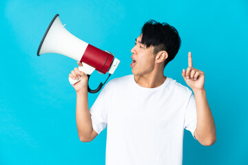 Young Chinese man isolated on blue background shouting through a megaphone to announce something in...
