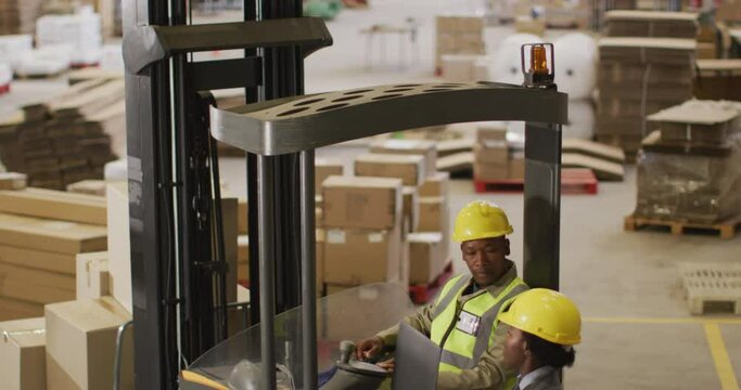 African american male and female workers wearing safety suits and using laptop in warehouse