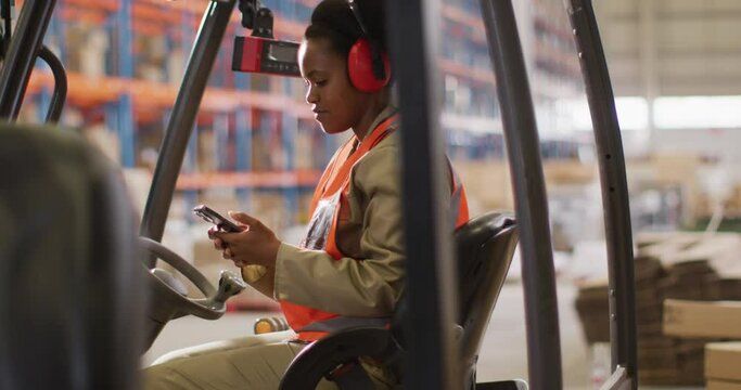 African american female worker wearing safety suit and using smartphone in warehouse