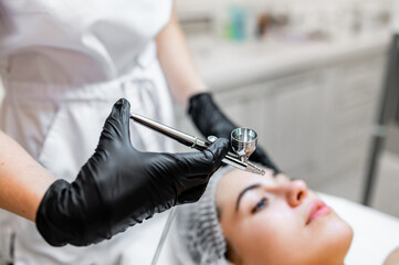 A hand in a black glove holds an airbrush against the background of the face of a pretty girl. Applying makeup with an airbrush. Hands in black latex gloves holding an airbrush. Services in the spa.
