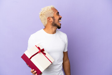 Young Colombian man holding a gift isolated on purple background laughing in lateral position