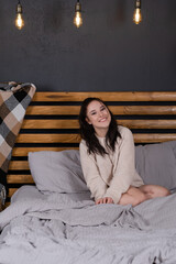 Fototapeta premium charming young woman in knitted beige sweater in bed with grey linens. modern loft bedroom in brown and gray colors