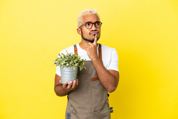 Young Colombian man holding a plant isolated on yellow background having doubts while looking up