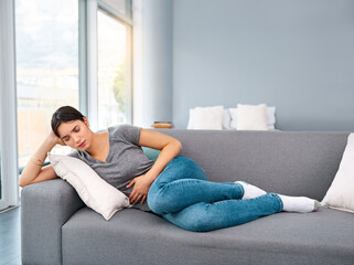 Obraz premium I think Im going to take easy today. Shot of a uncomfortable looking young woman holding a water bottle tightly while trying to get rest on a sofa at home.