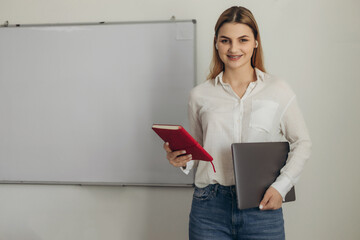 Portrait of a young teacher in the classroom. Girl holding a red notebook. Preparation for distance learning