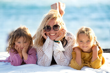 Fototapeta na wymiar Beautiful photo of cute happy little sisters playing with her mother outside. Woman and her daughters lying on the sunny beach. Happy family. Happy Mother's Day. Motherhood, childhood.