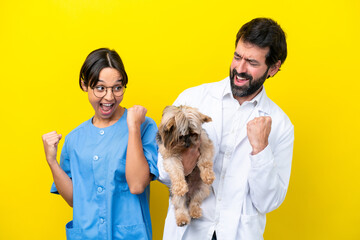 Young veterinarian couple with dog isolated on yellow background celebrating a victory