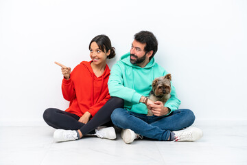 Fototapeta na wymiar Young caucasian couple sitting on the floor with their pet isolated on white background pointing to the side to present a product