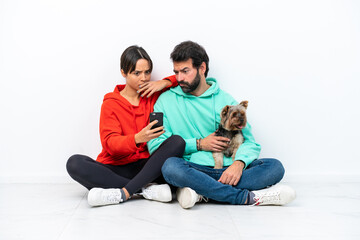 Fototapeta na wymiar Young caucasian couple sitting on the floor with their pet isolated on white background reading a message with the mobile