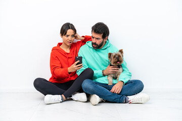 Young caucasian couple sitting on the floor with their pet isolated on white background reading a message with the mobile