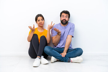 Fototapeta na wymiar Young caucasian couple sitting on the floor isolated on white background having doubts and with confuse face expression