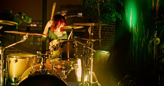 Cute drummer girl plays drums with drumsticks in recording studio with big round window and potted houseplants and flickering Concert RGB spot lights in slow motion. Camera moves from left to right.