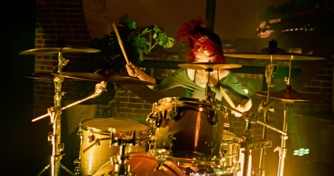 Cute drummer girl plays drums with drumsticks in recording studio with big round window and potted houseplants and flickering Concert RGB spot lights in slow motion. Camera moves from right to left.
