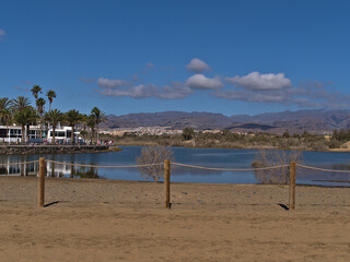 View of small lagoon La Charca in Meloneras, part of Maspalomas, southern Gran Canaria, Spain with fence in front and tourists walking on promenade.