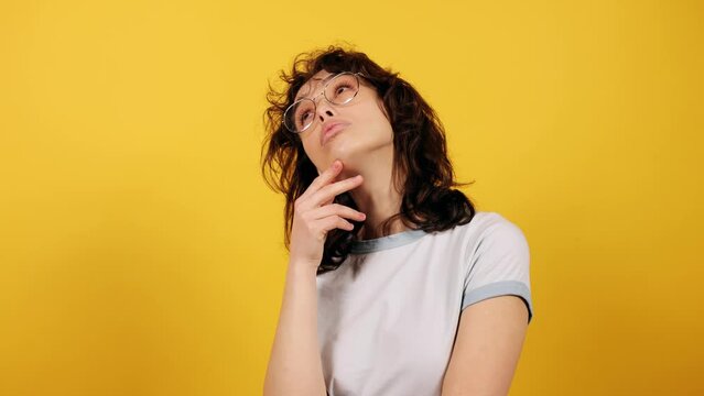 Young woman in eyeglasses thoughtfully pondering on camera. Attractive young woman on yellow background.