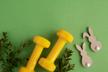 Two heavy dumbbells, decorative wooden Easter bunnies and boxwood branches. Healthy fitness lifestyle composition, gym workout and training concept. Fit flat lay with copy space on green background.