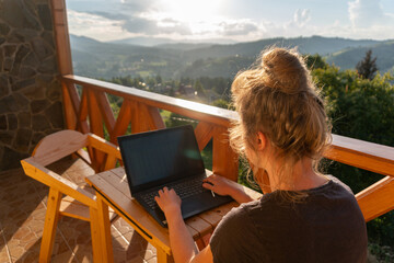 A young woman, freelancer is working on a laptop remotely on a balcony in the open fresh air near...