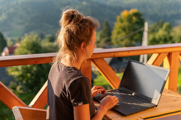 A young woman, freelancer is working on a laptop remotely on a balcony in the open fresh air near...