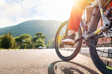Fototapeta na wymiar Cycling. A woman in red sportswear is sitting on a Bicycle with her foot on the pedal. Legs are close-up. Rear view of the road. Sunny. Concept of eco transport and healthy lifestyle