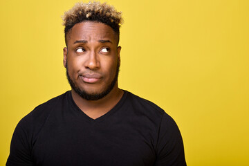 Fototapeta premium A dark-skinned man with a beard with a smirk on his face looks to the side in disapproval, faced with a difficult choice, dressed in an everyday black T-shirt. Doubting guy looks up with a smirk