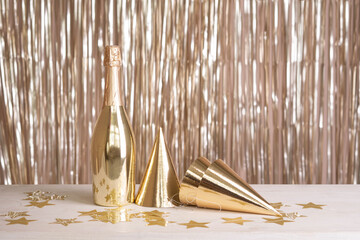 A bottle of champagne and party hats are on the table against the backdrop of a golden tinsel...