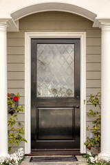 Front black door with decorative window. Two columns support the house. Red plants are on both sides of the entrance.