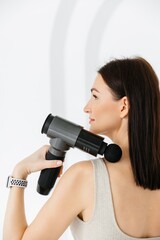 A girl at home massaging with a vibrating massager with a nozzle.  An electric therapeutic pistol massager in her hand massages the muscles of her arm. Sports recovery concept after a workout. 