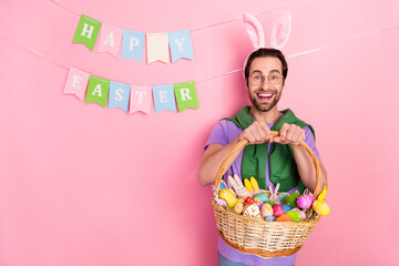 Photo of cheerful laughing childish man with rabbit ears present young easter gift isolated on pink...