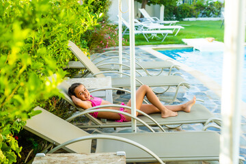 beautiful little girl resting on a sun lounger next to the pool against the backdrop of rows of sun...
