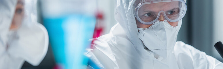 scientist in hazmat suit, goggles and medical mask near blurred colleague in lab, banner.