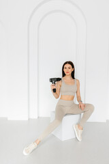 Fototapeta na wymiar A girl in a tracksuit is holding a vibrating massager. An electric pistol massager in her hand massaging the muscles. Sports recovery concept after a workout. 