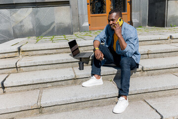 Fototapeta na wymiar Young attractive african american freelancer businessman or student sitting on stairs in urban area and doing some work on laptop. Handsome male take break and drink take away coffee