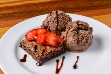 Brownie with sliced strawberries and chocolate ice cream on a white plate on a wooden table.