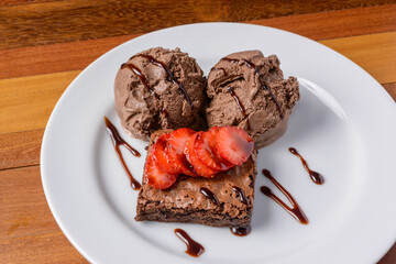 Brownie with sliced strawberries and chocolate ice cream on a white plate on a wooden table.