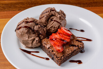 Brownie with sliced strawberries and chocolate ice cream on a white plate on a wooden table.
