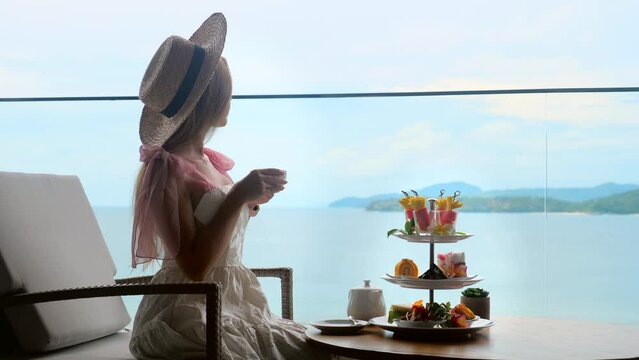 Young lady in white dress drinking tea on Afternoon tea with mini canape and selection of various sweets with sea on background. Elegant woman enjoy ocean view, eating desserts in luxury restaurant.