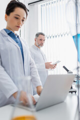 Fototapeta premium young scientist using laptop near mature colleague with digital tablet on blurred foreground.