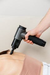 A therapist massages a woman's back with a massage percussion device in her home. The therapist's hand holds a therapeutic vibrating massager. Physical therapy and muscle recovery and massage