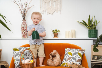 A little smiling boy standing on the orange sofa  with easter bunny, watering can and basket with...