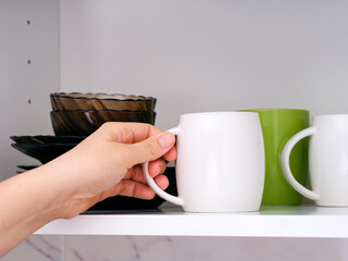 Woman hand taking a white mug from a kitchen shelf.