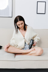 A girl at home on the couch massaging with a vibrating massager.  An electric therapeutic pistol massager in her hand massages the muscles of the foot. Sports recovery concept after a workout. 