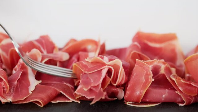 Prosciutto crudo or jamon meat on serving board on white background. Hand with fork takes Prosciutto. Sliced into thin pieces of ham on cutting serving  board. Italian antipasto 