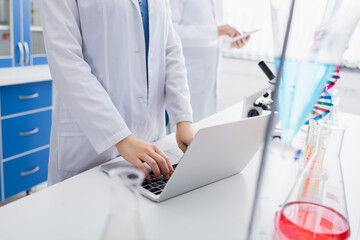 cropped view of scientist typing on laptop in laboratory near blurred colleague.