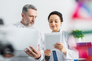 smiling scientist showing digital tablet to mature colleague with laptop.