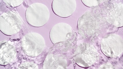 Poured water splashing and making ripples over cotton pads arranged in rows on purple background | product background, cleansing lotion commercial