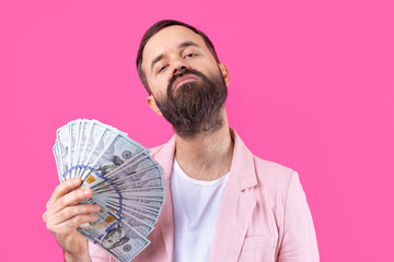 Portrait of a contented young businessman with a beard dressed in a pink jacket showing us dollar banknotes against a red studio background. Taste, smell of money.